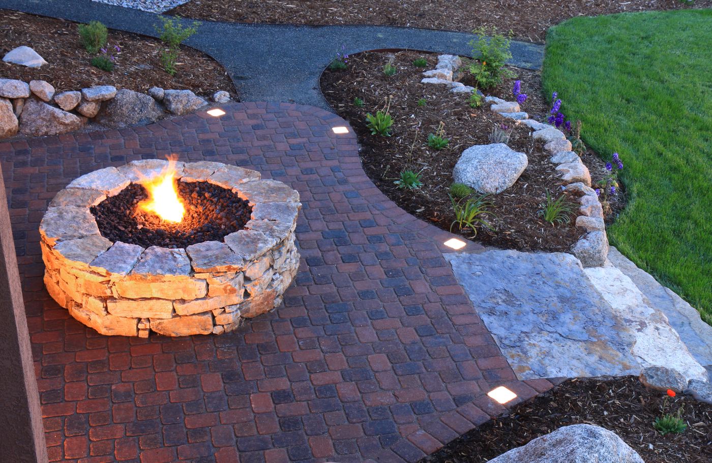 A curved paver patio surrounding a stone fire pit and landscaped garden.